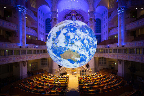 GAIA in der Frauenkirche: Foto einer beleuchteten Erdkugel, die frei in einem Kirchenraum hängt. Darunter sind Kirchenbänke und Personen, dahinter Altar und Orgel zu sehen.