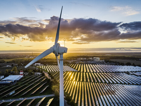 Foto von einem Windrad vor untergehender Sonne, dahinter ist eine großflächige Solaranlage zu sehen