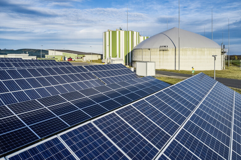 Großflächige Photovoltaikanlage mit sonnenenergieerzeugenden Solarmodulen im Vordergrund. Im Hintergrund sind zwei große, kuppelartige Biogaslager sowie technische Gebäude und Anlagenkomponenten zu erkennen.