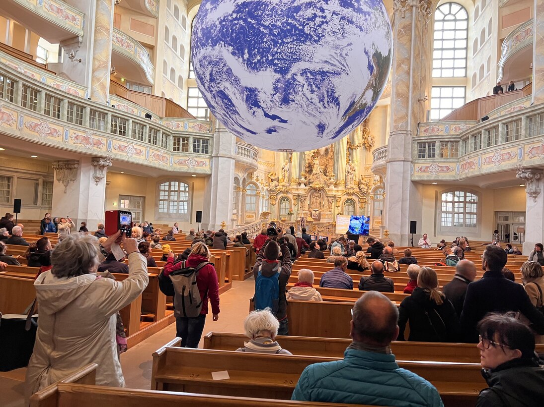 Blick in den Kirchenraum der Frauenkirche Dresden: Die Kunstinstallation GAIA schwebt über den Kirchenbänken. Auf den Bänken sitzen zahlreiche Personen.