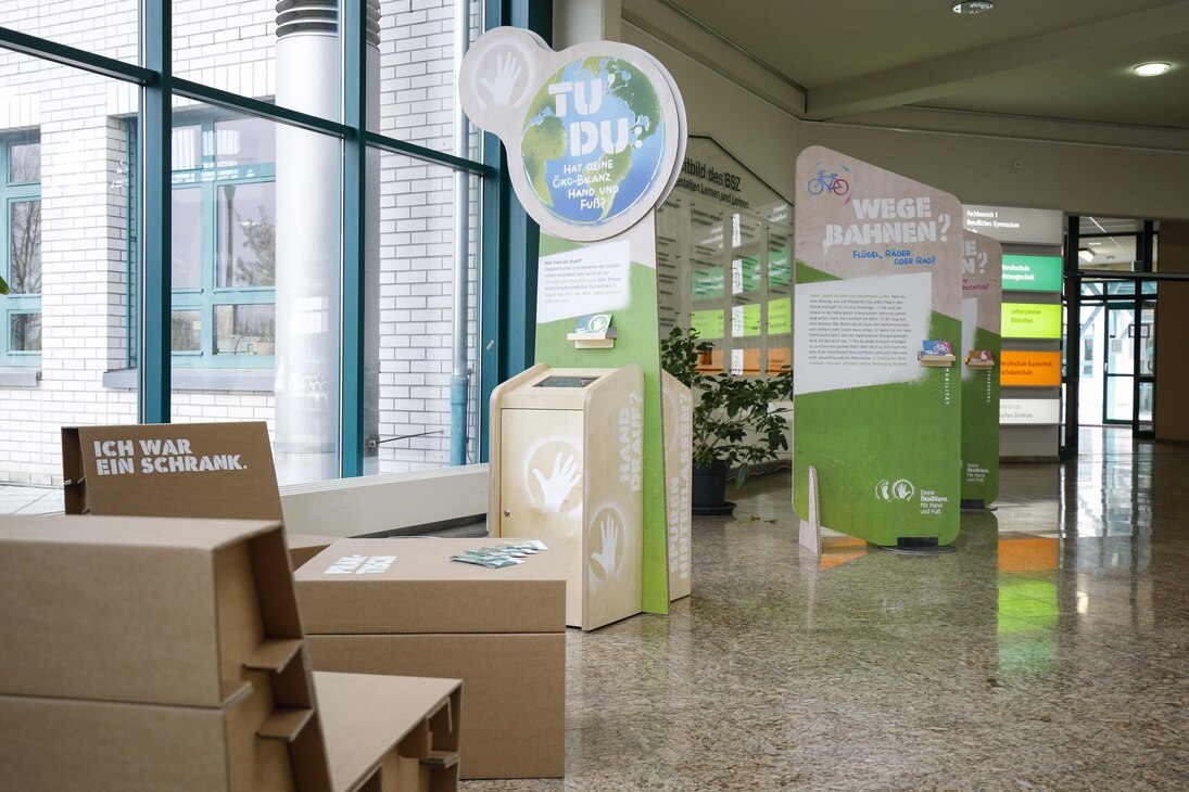 Foto der Ausstellung »Hat Deine Ökobilanz Hand und Fuß?« in einem Schulgebäude. Im Vordergrund stehen Möbel aus Pappe, im Hintergrund sind die Info-Aufsteller der Ausstellung zu sehen. 