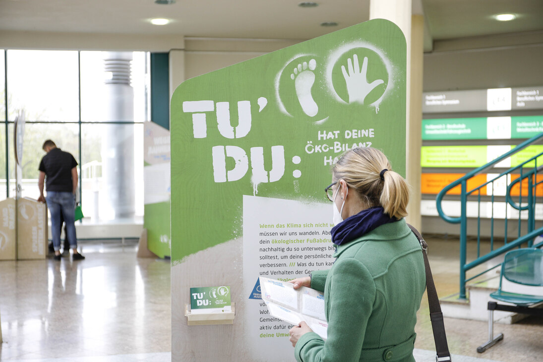 Foto der Ausstellung »Hat Deine Ökobilanz Hand und Fuß?«: Eine Person mit grüner Jacke steht vor einem Aufsteller und liest einen Info-Flyer.