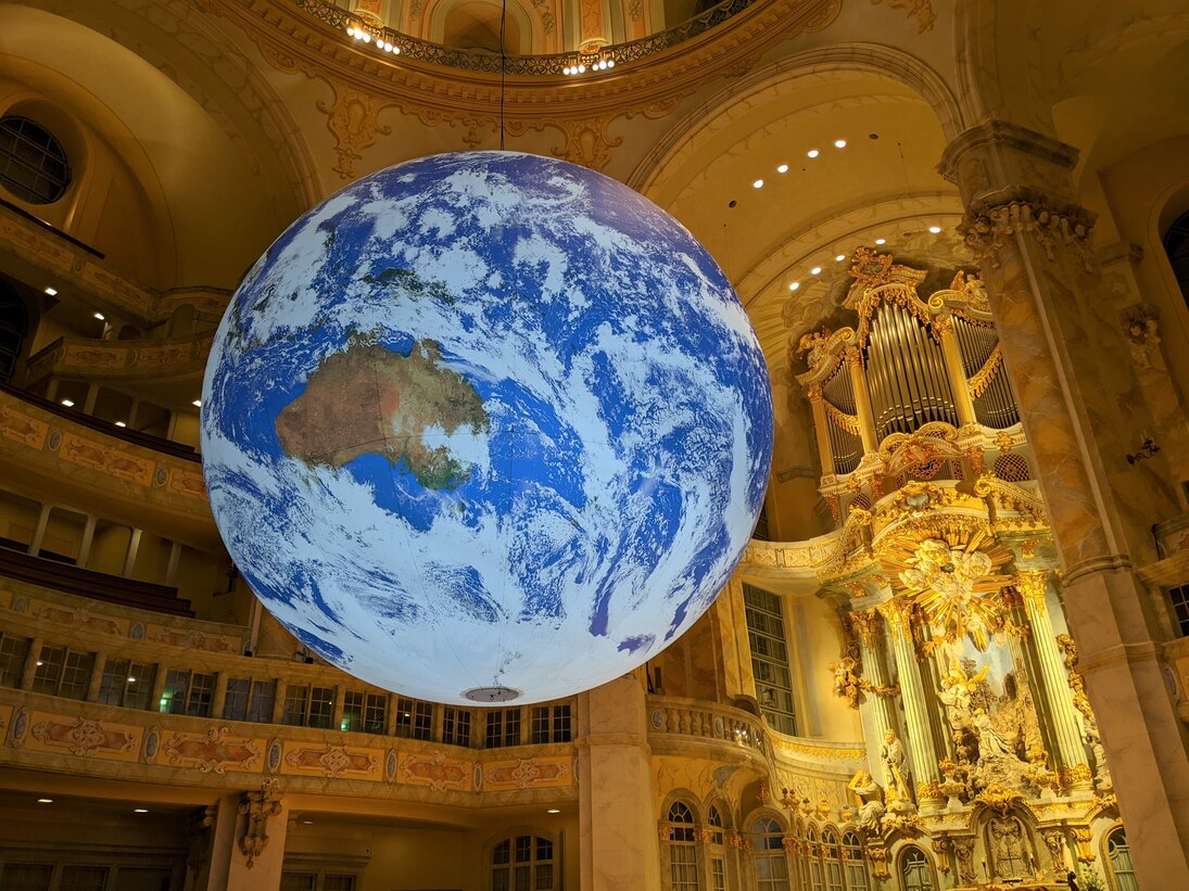 Foto der Kunstinstallation »Gaia«: eine beleuchtete Erdkugel hängt frei im Kirchenraum der Dresdner Frauenkirche, dahinter sind Emporen und der beleuchtete Altar zu sehen 