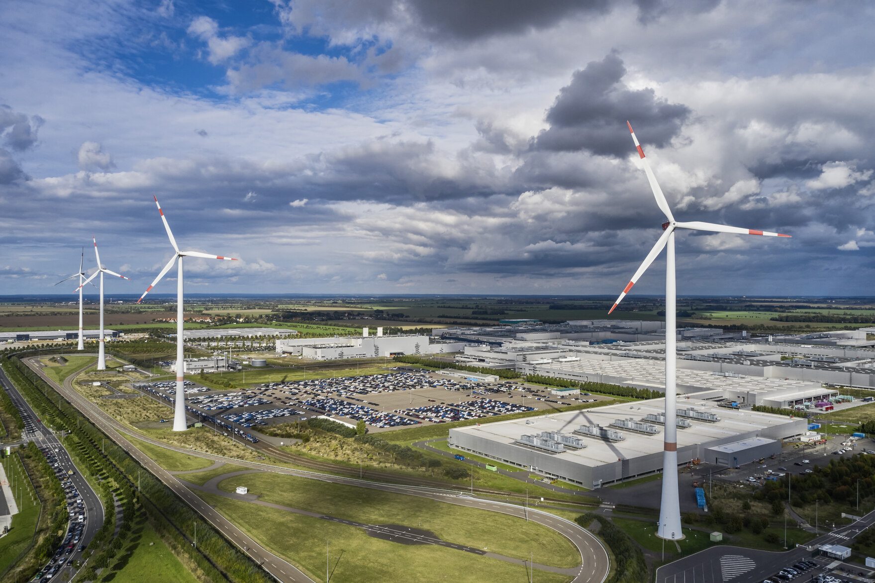 Foto von vier Windrädern am BMW-Werk Leipzig. Hinter den Windrädern sieht man Industrie-Gebäude und einen bewölkten Himmel.