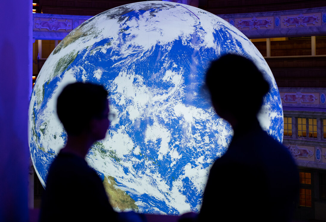 Foto der Kunstinstallation »Gaia«: eine beleuchtete Erdkugel hängt im Kirchenraum der Dresdner Frauenkirche, davor sind die Silhouetten von zwei Personen zu sehen.