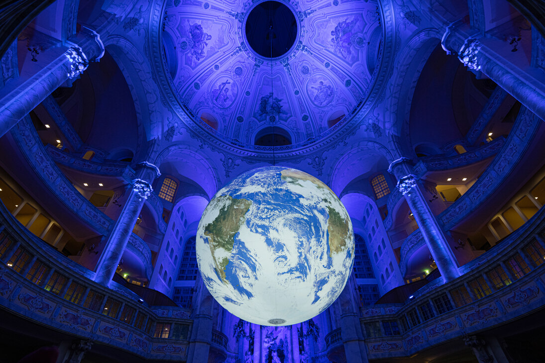 Foto der Kunstinstallation »Gaia«: eine beleuchtete Erdkugel hängt frei im Kirchenraum der Dresdner Frauenkirche, dahinter sind die Emporen und die Kuppel zu sehen. Der Kirchenraum ist blau beleuchtet.