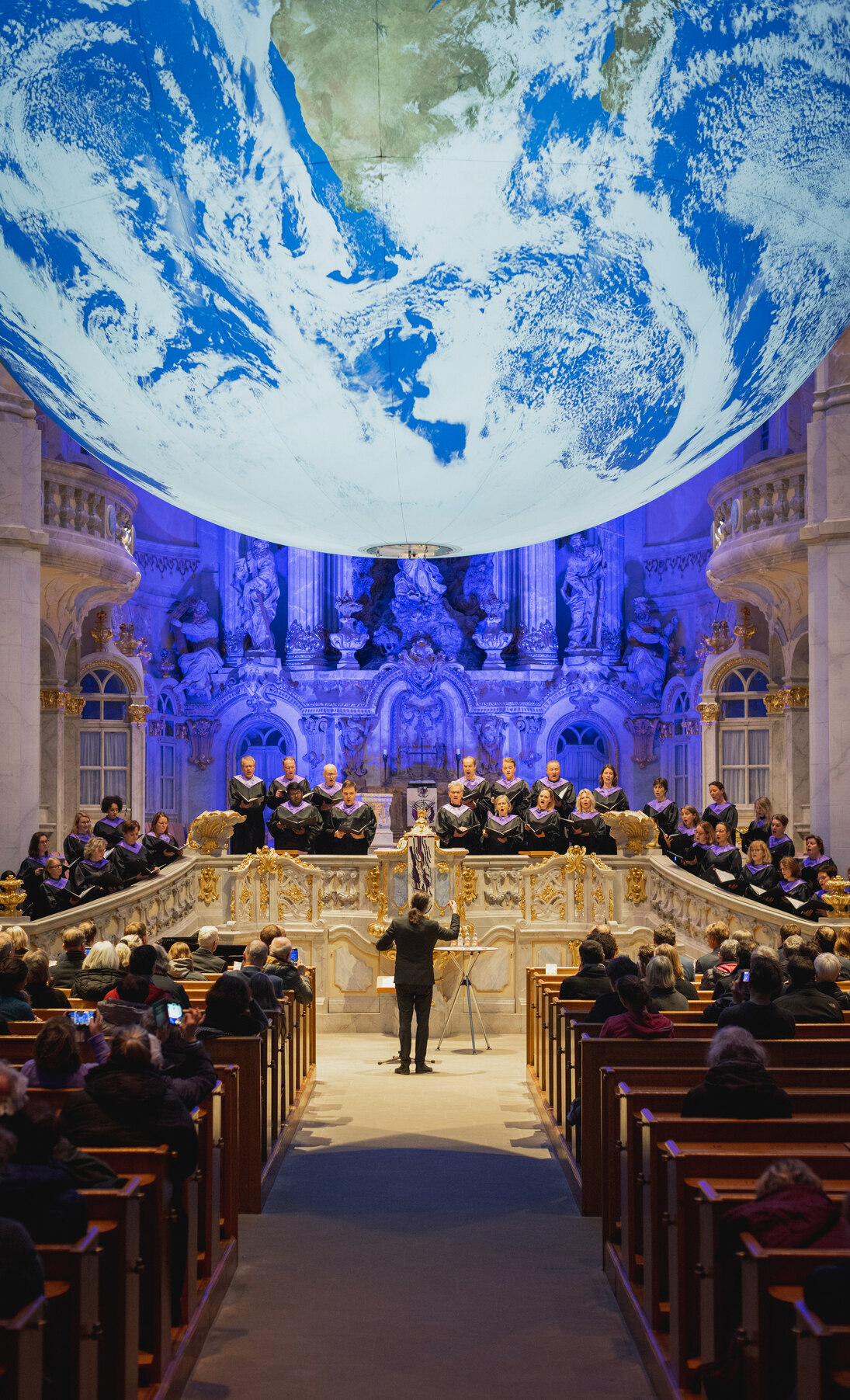 Foto von der Eröffnung zur Kunstinstallation »Gaia«: Auftritt vom Chor der Frauenkirche Dresden, darüber schwebt die beleuchtete Erdkugel »Gaia«, davor sieht man Kirchenbänke mit Besuchern.